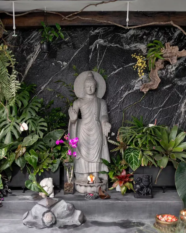 A tranquil corner in the 'Sun Room' project. A stone Buddha statue is surrounded by lush tropical foliage like Monstera and Philodendron, with mounted orchids and ferns against a black marble wall, creating a space for peace.