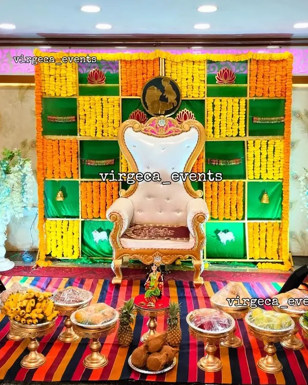 A vibrant Seemantha decoration featuring a traditional checkerboard backdrop of green, yellow, and orange marigolds. The setup is completed with an ornate chair and a full display of seer plates for the ceremony.