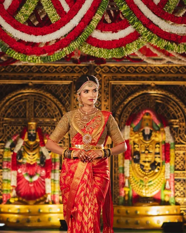 A bride standing before a magnificent temple-style mandap. The background features golden idols and is framed by rich red and white flower garlands, creating a divine atmosphere.