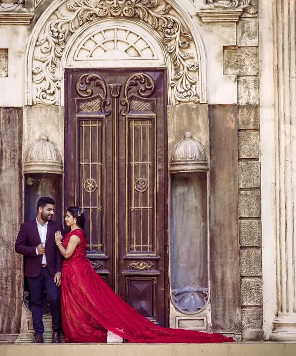 Create a timeless pre-wedding portrait with this stunning maroon-red floral gown. Paired with a classic men's blazer, it makes for a powerful and romantic couple's look against a grand architectural backdrop.