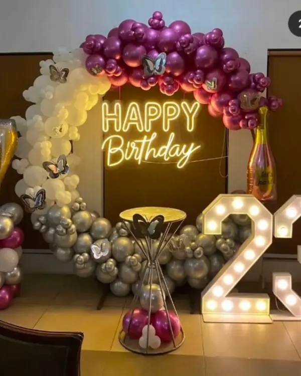 A complete party setup for a 23rd birthday. It features a large circular balloon arch in purple, white, and silver, a custom "Happy Birthday" neon sign, and big light-up marquee numbers.