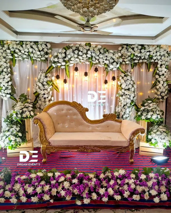 An elegant engagement or reception stage featuring a beautiful arch of white flowers, a classic chaise lounge, and warm accent lighting for a timeless look.