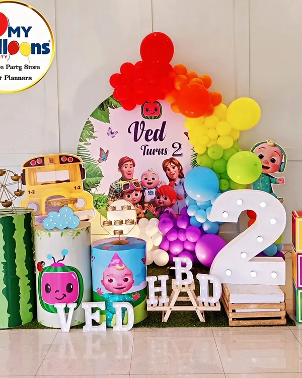 A complete Cocomelon-themed second birthday setup. This vibrant decor includes a rainbow balloon arch, the iconic yellow school bus, character-printed plinths, and a large light-up number '2'.