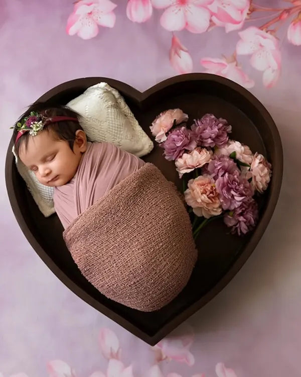 A peaceful newborn rests in a heart-shaped wooden bowl, surrounded by a soft pink floral background. This image showcases my styled newborn sessions, where I use props and safe posing to create artistic portraits.