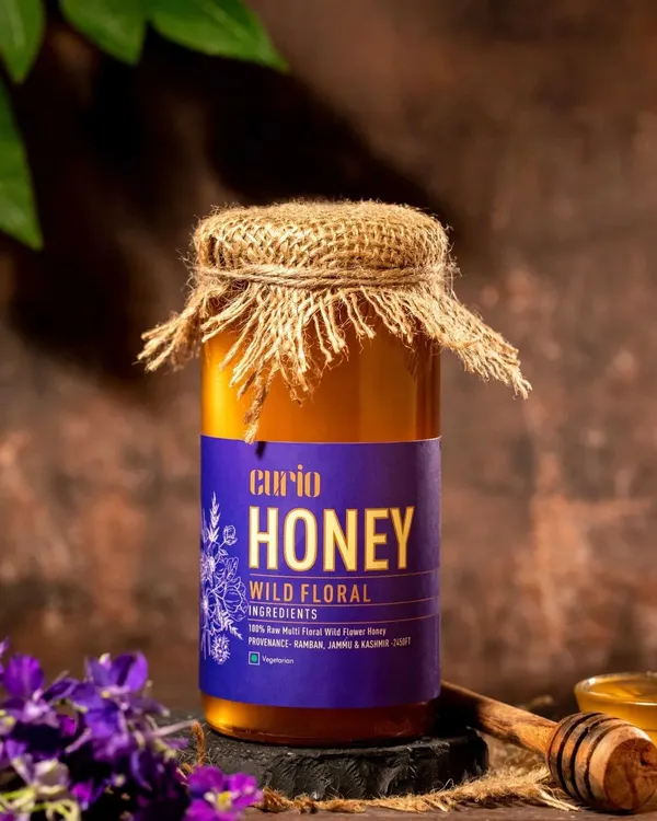 This image of wild floral honey is styled to feel rustic and authentic. The jute-covered lid, dark background, and purple flowers hint at the honey's natural origins and unique flavor profile.