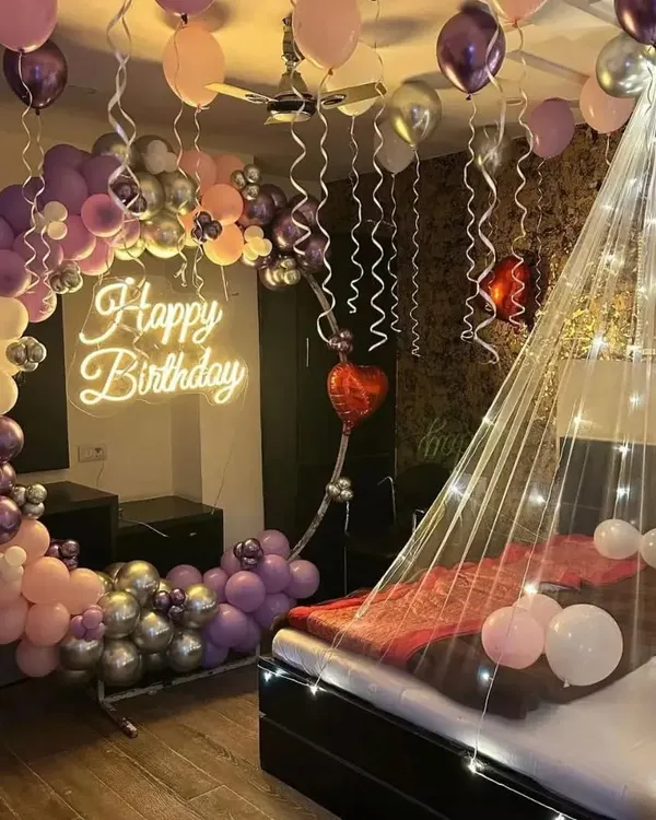 A romantic birthday surprise in the bedroom. This setup features a bed canopy with fairy lights, a half-arch of balloons in purple and pink, and a "Happy Birthday" neon sign.