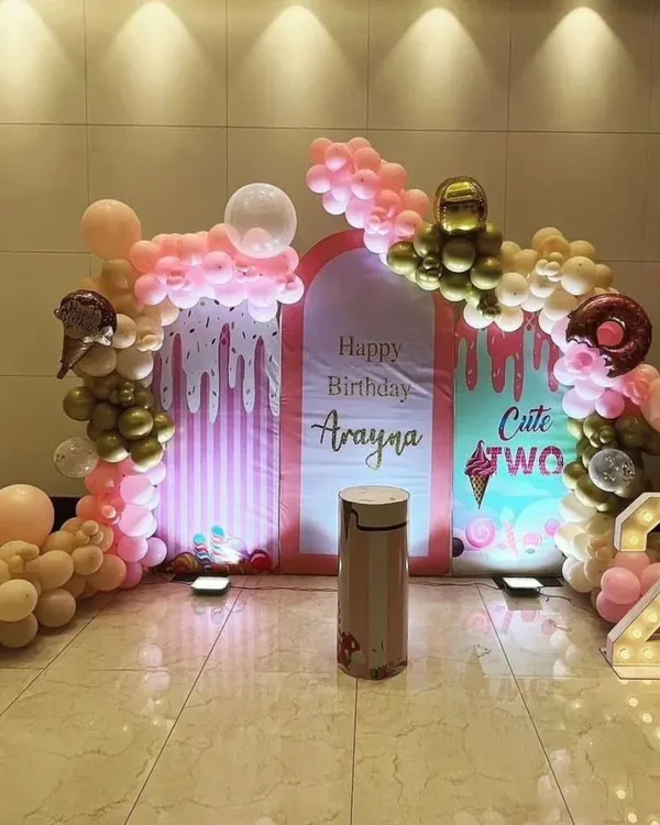 A "Cute Two" ice cream theme for a second birthday. The setup includes pastel pink and gold balloons and backdrops with dripping ice cream patterns.