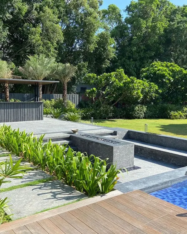 A modern farmhouse landscape featuring a sunken seating area with an integrated fire pit adjacent to the pool. This design creates distinct zones for relaxation and entertainment, surrounded by lush plantings and an outdoor bar for a complete resort-like experience at home.