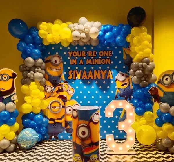 A complete Minions theme party setup for a 3rd birthday. This vibrant decoration features a "You're One in a Minion" backdrop, character cutouts, a light-up number '3', and balloon columns in the signature yellow and blue.