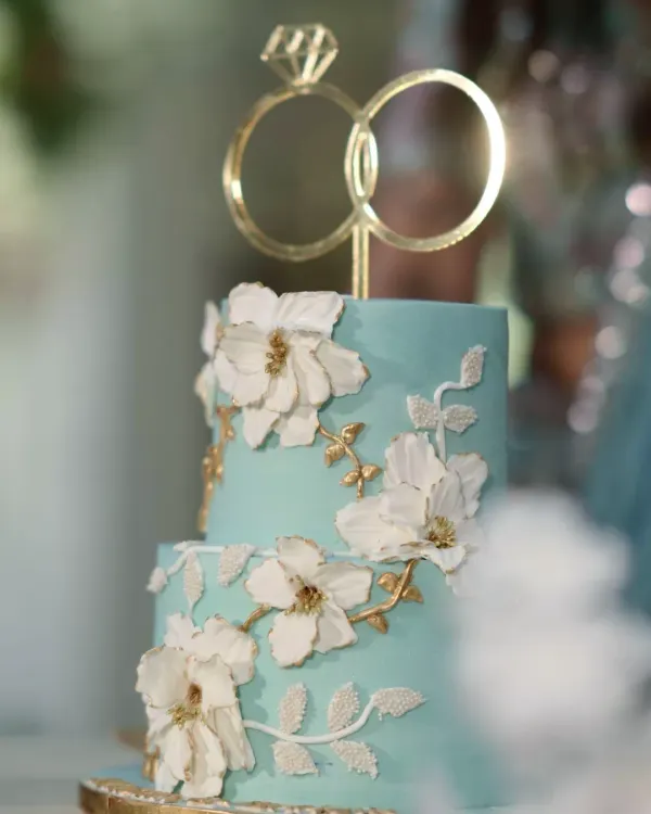 A close-up of this dreamy two-tiered engagement cake. The soft blue ganache is the perfect canvas for the handcrafted white and gold sugar flowers, topped with elegant interlocking rings.