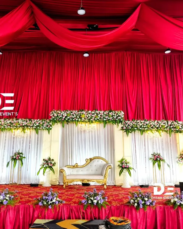 A classic reception stage featuring bold red ceiling drapery that creates a dramatic effect. The backdrop combines clean white panels with tiers of fresh flowers, perfectly framing the elegant couple's sofa.