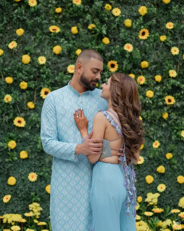 A sweet moment between a couple against a vibrant wall of yellow flowers during their Haldi ceremony. Their coordinated pastel blue outfits create a beautiful contrast, making for a fresh and modern portrait.