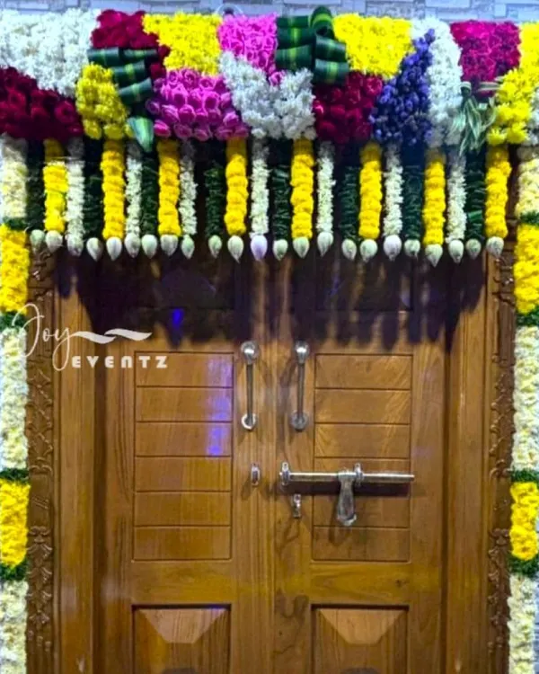 An elaborate main door flower decoration for a housewarming ceremony. This design features a dense frame of multi-colored flowers and jasmine buds, creating a grand and festive entrance.