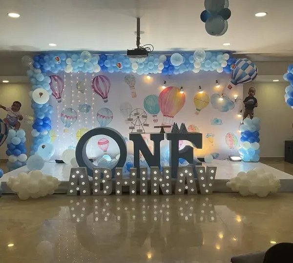 A spectacular hot air balloon themed stage for a first birthday. The grand setup includes marquee lights, multiple balloon pillars, and a wide backdrop depicting a sky full of hot air balloons.
