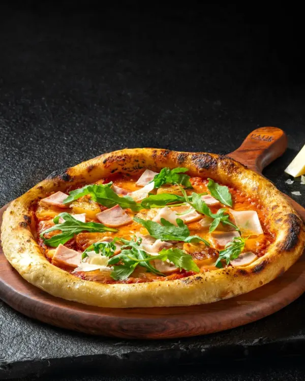 A classic ham and arugula pizza on a wooden board, shot with dramatic side lighting to highlight the crispy crust and fresh toppings.