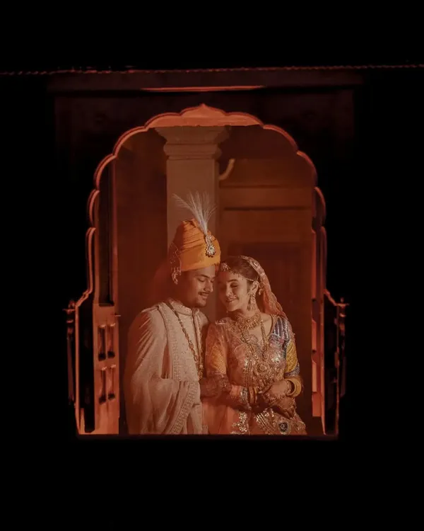 A cinematic, fairytale-like shot of the couple framed within a traditional Rajasthani jharokha window, creating a private and romantic moment.