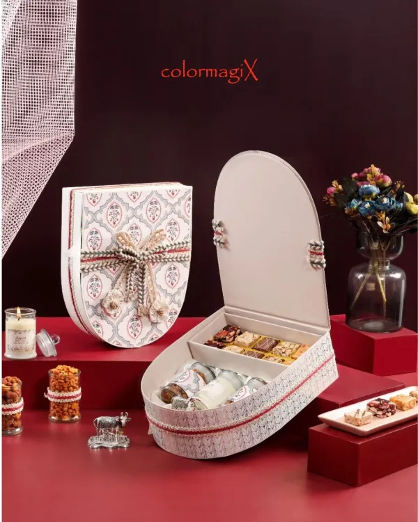 A uniquely shaped shield-style gift box with a traditional Indian motif and an elaborate woven ribbon. The box opens to reveal compartments filled with luxury sweets.