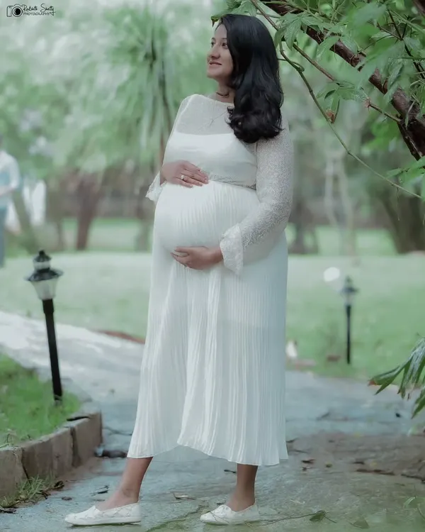 A full-length portrait of the mother-to-be in a flowing white dress, looking thoughtfully into the distance. The natural setting complements her serene expression.