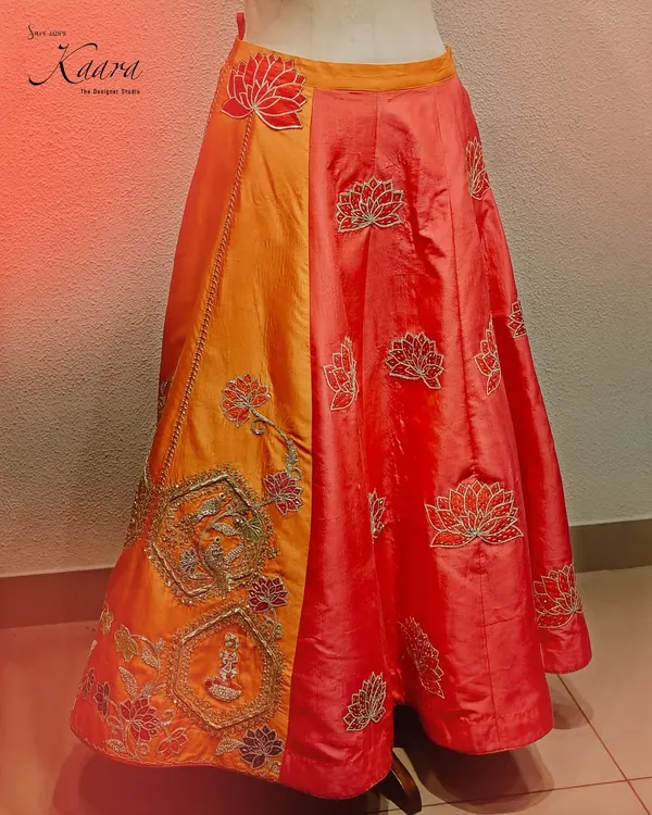 A celebration of vibrant color and traditional motifs. This lehenga features a rich orange panel with intricate zardosi work depicting a wedding scene, contrasted with a red panel adorned with delicate lotus embroidery.