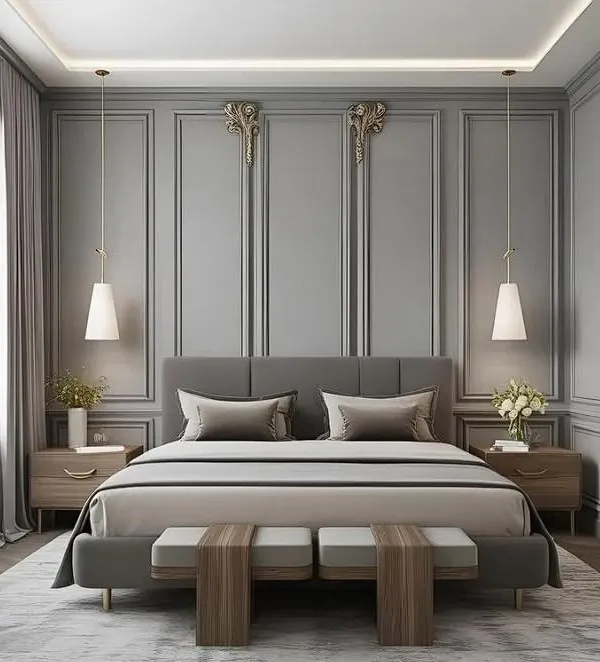 A symmetrical and elegant bedroom design featuring a grey upholstered bed set against a wall with classic panel molding. Matching pendant lights hang over wooden nightstands, and a pair of benches at the foot of the bed completes the sophisticated look.