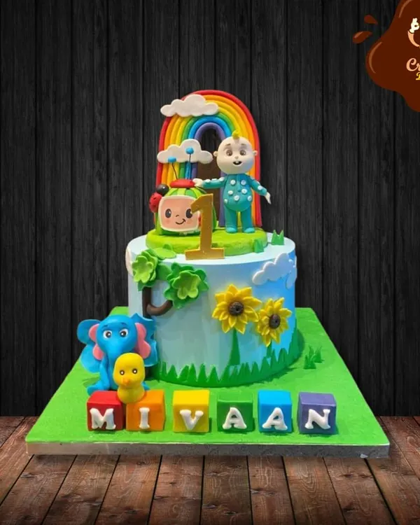 A bright and cheerful Cocomelon cake for a first birthday. This single-tier cake features a rainbow, clouds, and characters from the show, with the birthday boy's name spelled out in colorful blocks.