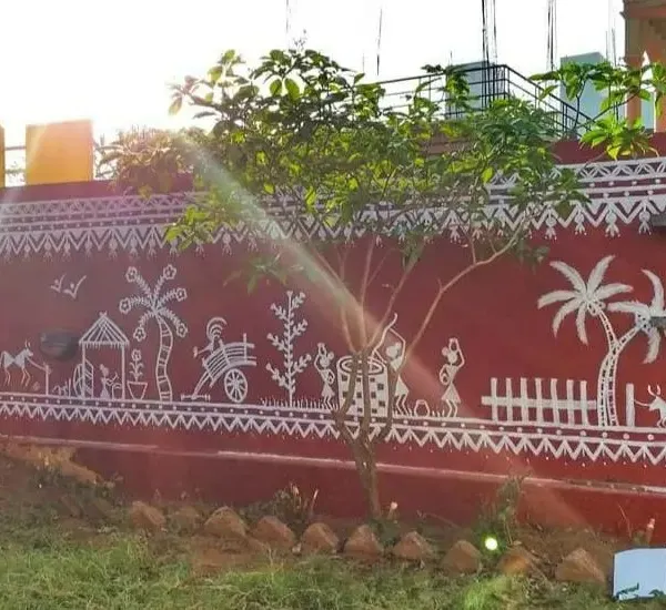 A wide view of the completed Warli mural, showcasing a detailed scene of rural life. This traditional art form, painted on a striking red exterior wall, tells a continuous story featuring people, animals, and nature.