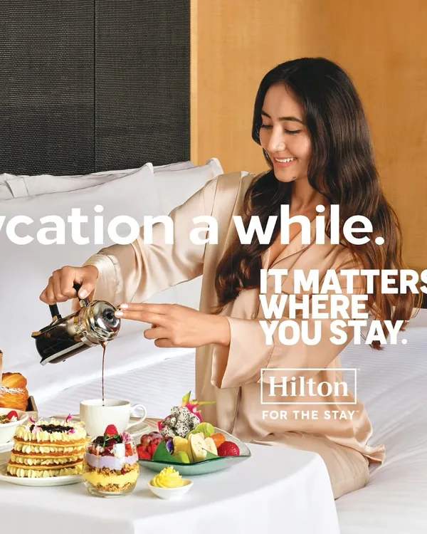 A serene moment styled for a Hilton Hotels campaign. The model's luxurious silk pajamas and the beautifully arranged breakfast tray create an aspirational 'staycation' scene, emphasizing comfort and indulgence.