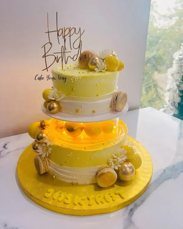 This two-tiered cake was designed for a special birthday, featuring a beautiful yellow and white theme. It's illuminated from within for a magical glow and adorned with golden macarons and delicate sugar flowers.