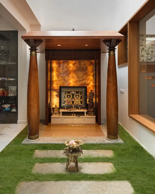 A frontal view of the pooja space in the Twin Courtyard House. The mandap is framed by two large, reclaimed wooden pillars, and the backlit onyx panel creates a divine glow.