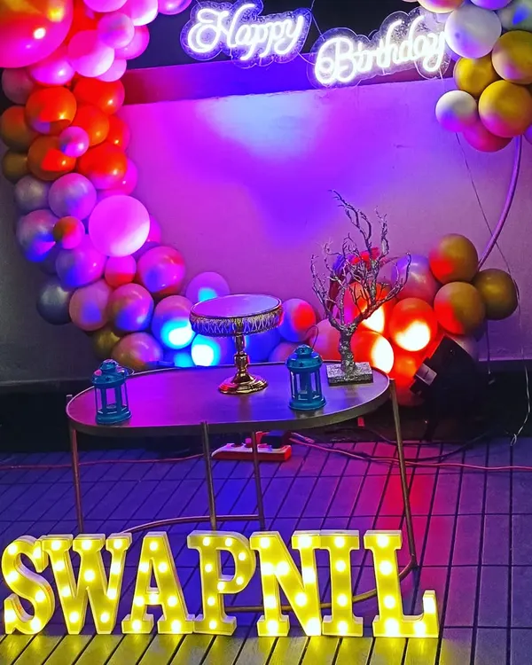 A surprise birthday party for a husband, featuring a modern ring circle balloon decoration. The setup includes a neon "Happy Birthday" sign, marquee letters, and a mix of pastel and chrome balloons for a stylish look.
