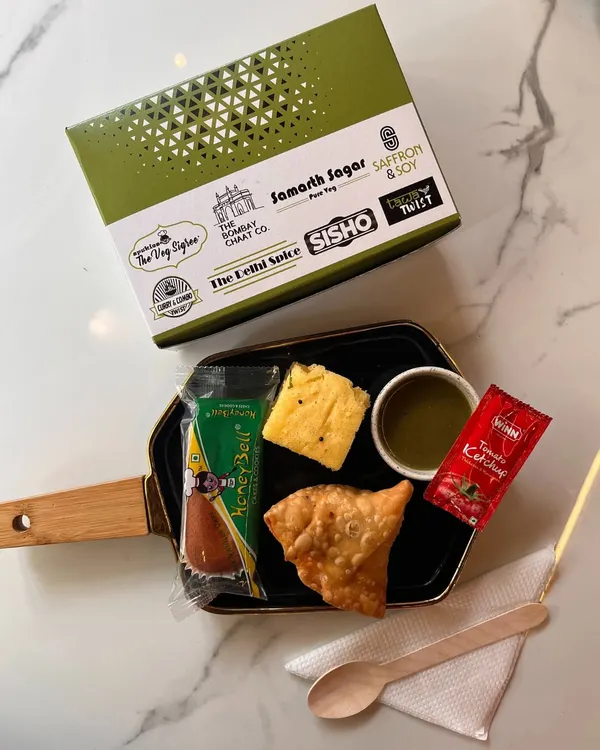 Our standard snack box is designed for convenience and taste. This one features a classic combination of a crispy samosa, soft dhokla, a sweet honey bell cake, and mint chutney, all packed in our branded, eco-friendly box.