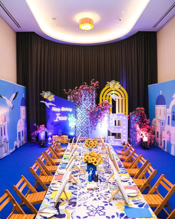 A stunning Greece-themed art party setup, with a long table ready for canvas painting, set against a backdrop of Santorini-style buildings and beautiful bougainvillea flowers.