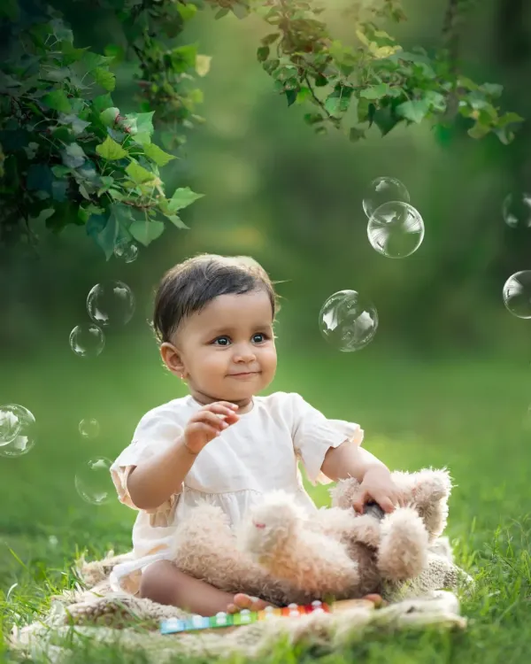 A baby sits on a blanket in a lush green park, mesmerized by floating bubbles. An outdoor sitter session like this is perfect for capturing natural wonder.