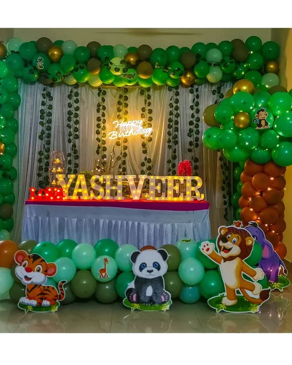 A full jungle theme decoration perfect for a living room setup. This design features a lush green balloon arch with hanging vines, a balloon tree, a neon sign, marquee name lighting, and friendly animal cutouts like a panda, tiger, and lion.