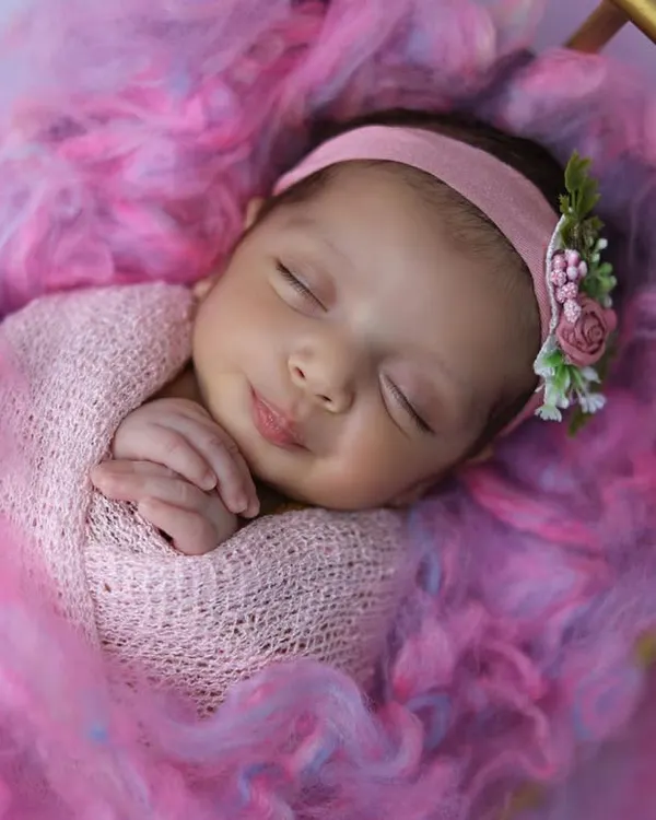 A perfect sleepy smile on a baby girl, swaddled in pink and nestled in a bed of soft, colorful wool. This is a classic, heartwarming newborn portrait that parents adore.