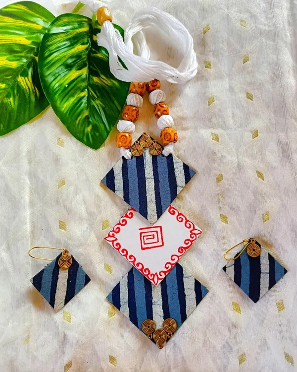 This geometric fabric jewelry set plays with shapes and patterns. The diamond-shaped pendant combines striped blue fabric with a hand-painted central element. The matching earrings and beaded cord complete this modern, lightweight accessory.
