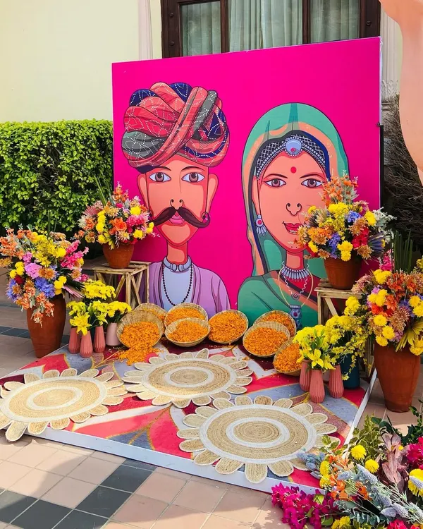A quirky and colorful Mehendi photo booth with a custom Rajasthani-style caricature backdrop and vibrant floral arrangements.