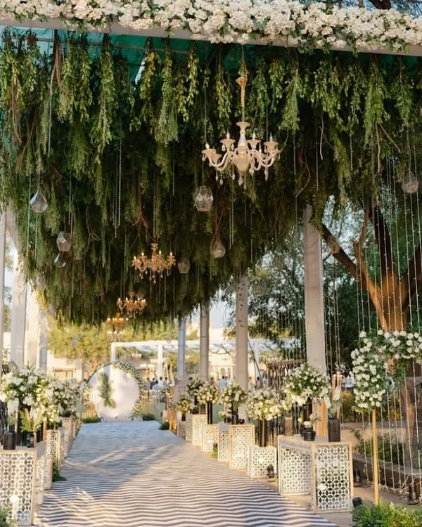 A truly grand and elegant wedding entrance. The walkway features a ceiling of hanging greenery and chandeliers, with the path lined by intricate white pedestals and floral arrangements.