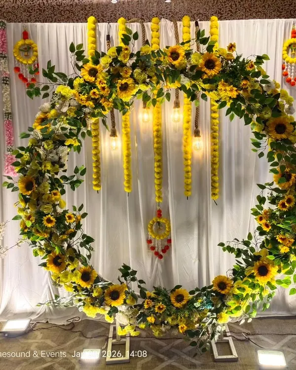 This modern Haldi ceremony backdrop features a full ring of bright sunflowers and green foliage. We've added hanging marigold garlands and vintage-style bulbs to create a perfect photo spot for your event.