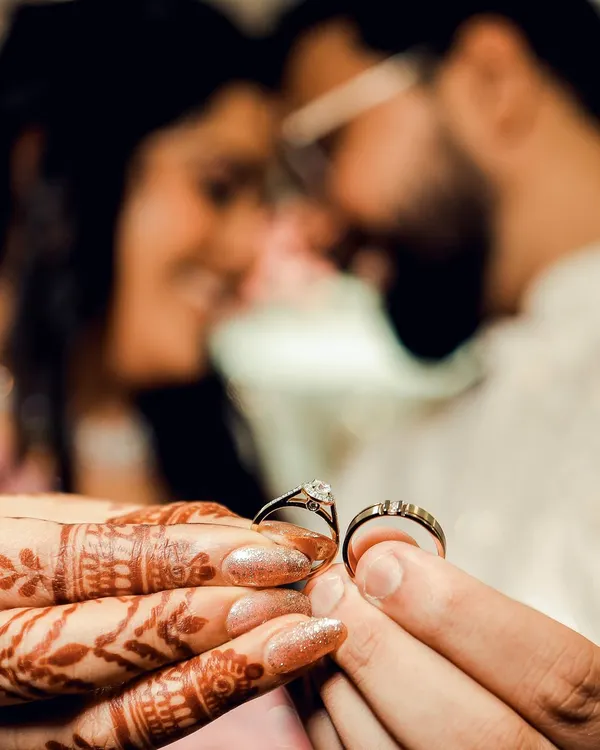 A creative shot of the wedding rings held by the couple. The focus is on the rings, with the couple blurred lovingly in the background, symbolizing that their union is at the center of it all.
