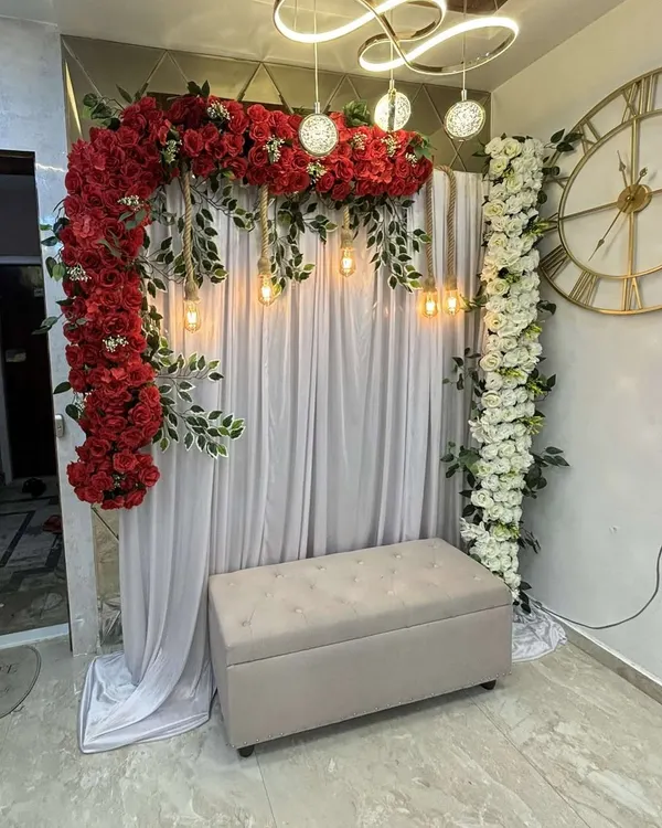 A beautiful and modern engagement photo backdrop. This setup features a lush arch of red roses against a clean white curtain, with hanging vintage bulbs for a warm glow. A comfortable bench provides the perfect spot for couple and guest photos.