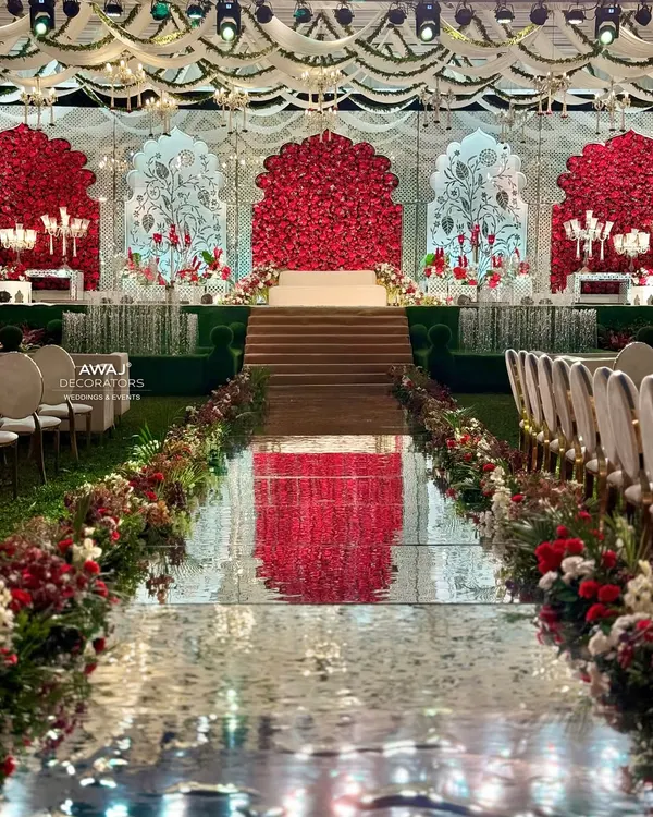 A breathtaking view of a grand reception stage, where a mirrored aisle reflects the magnificent red and white floral backdrop, creating a path that feels like walking on air towards your new beginning.