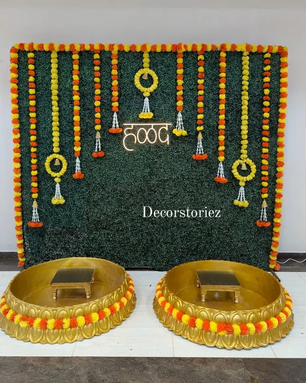 We designed this complete Haldi ceremony setup featuring a lush artificial grass wall backdrop, traditional marigold garlands, and a bright neon 'हळद' sign. The two large golden Urlis and matching patlas (stools) are also available from us on a rental basis.