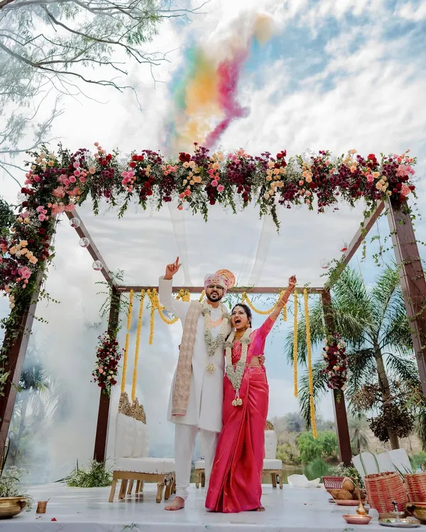 A triumphant moment for the couple under a shower of colorful smoke. This shot captures the celebratory climax of the wedding ceremony, framed by a beautiful floral mandap.