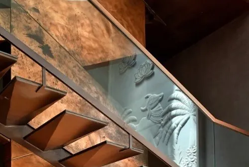 This copper and metal staircase is a study in lightness and intensity. The cantilevered treads seem to float, while the glass balustrade offers a glimpse of the traditional 'pichwai' relief art on the wall, lit by a skylight above.
