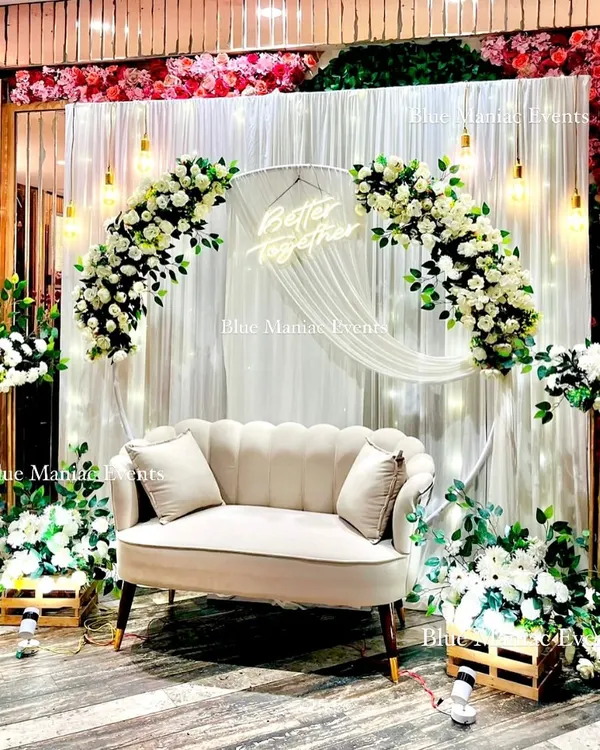 I specialize in creating stunning backdrops for your most cherished moments. This elegant Roka setup features a full circular frame wrapped in white florals, set against a fairy-lit curtain and a 'Better Together' neon sign, creating a perfect photo opportunity.