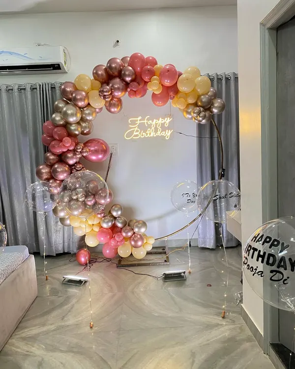 An elegant balloon ring decoration for an intimate home celebration. This setup features a half-arch of chrome and pastel balloons on a golden ring, with a 'Happy Birthday' neon sign.