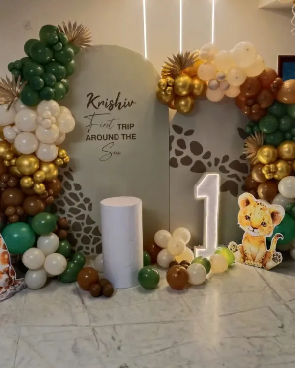 This jungle safari theme is one of my most requested setups for a first birthday. It features a custom two-panel backdrop with the baby's name, a large light-up number '1', and organic balloon garlands in earthy green, white, and gold tones, complete with cute animal cutouts like a giraffe and lion cub.