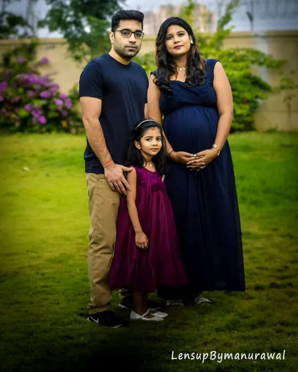 An expecting couple and their daughter pose for a beautiful outdoor maternity shoot in a lush green garden, capturing the family's anticipation in a natural setting.