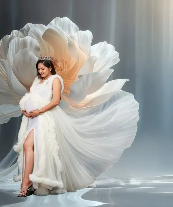 A serene and artistic portrait of a mother-to-be in a flowing white gown, cradling her bump. She is surrounded by a giant, blooming fabric flower, creating a dreamy, ethereal atmosphere that celebrates the beauty of new life.